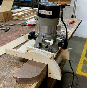 Image of a light wood jig, metal router holding a curved piece of dark brown wood on a large work table.