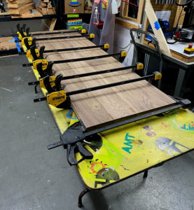 Image of several long pieces of wood held in 6 horizontal clamps on a yellow folding table