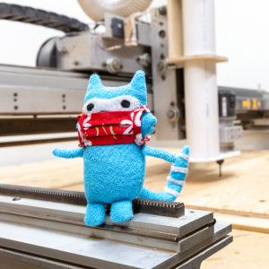 Blue Plushie with Red Mask sitting on the tracks of a CNC Router