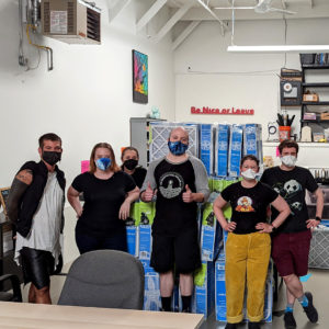 Image of 6 volunteers, in wearing black with mask stand in front of an apx. 6ft stack of blue DIY Filter Kits