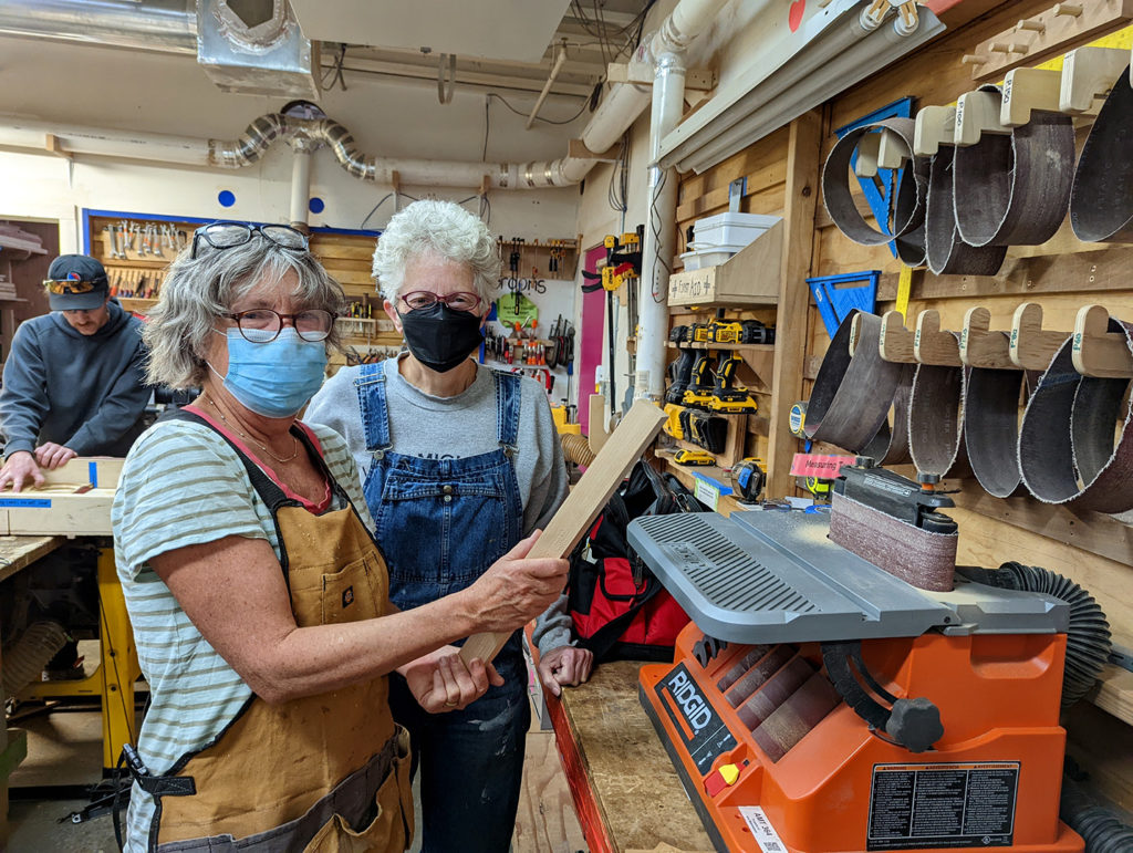 Two folks posing with a piece of wood next to a sander.