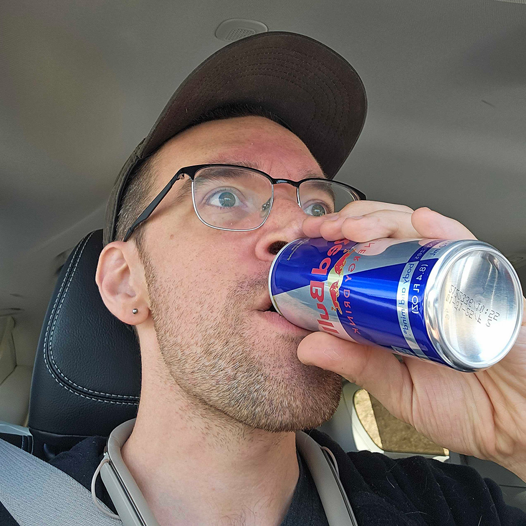 Person in baseball cap drinking Red Bull in car.