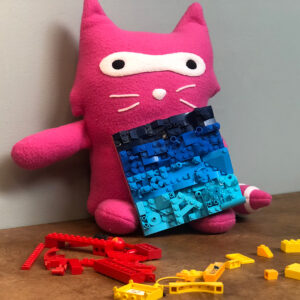 stuffed pink raccoon with a blue lego mosaic and scattered yellow and red logo bricks. The critter looks mildly drunk.