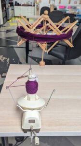 A pull skein of purple yarn being wound on a ball winder from a hank draped over a wooden swift.