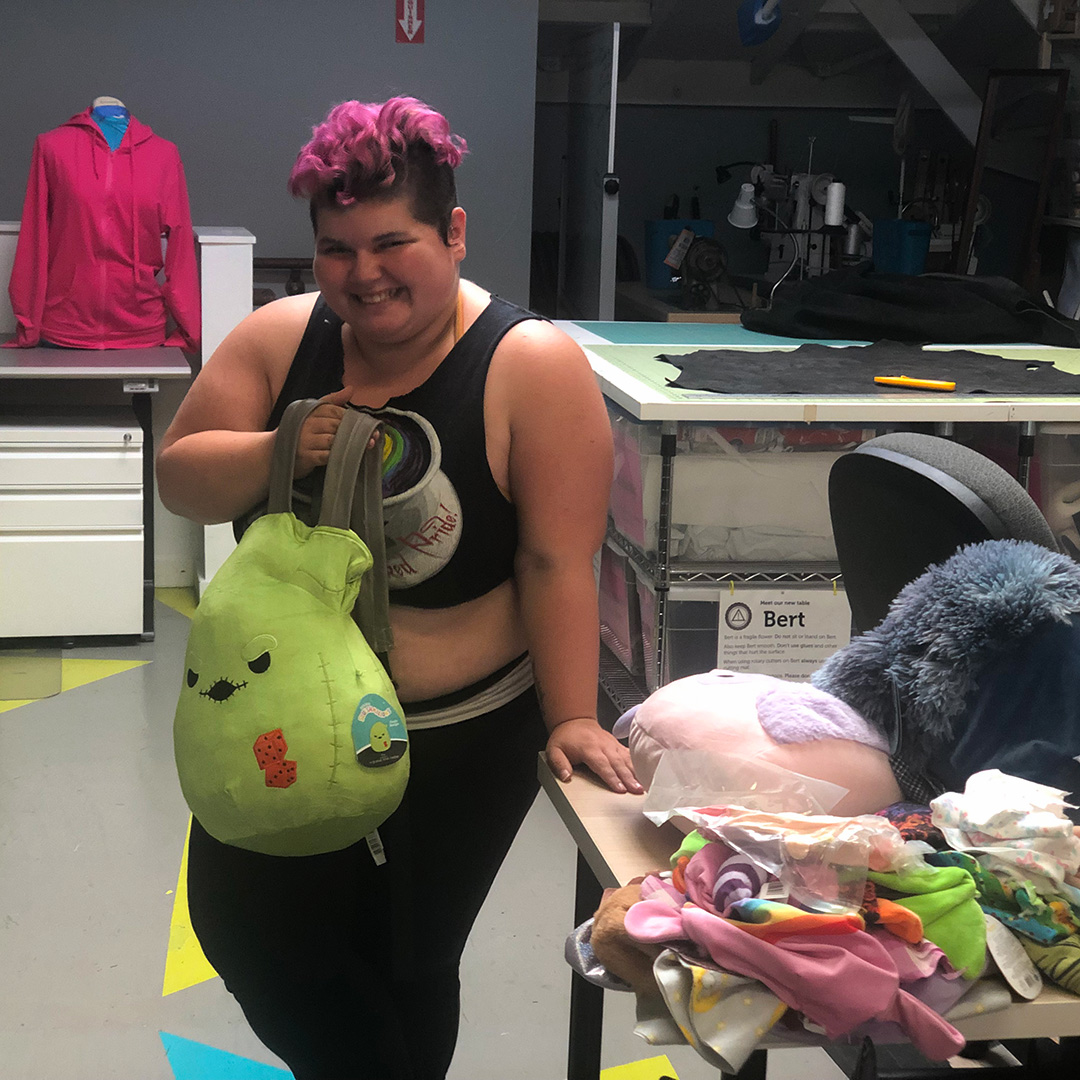 Person with pink hair holding green Squishmallow backpack
