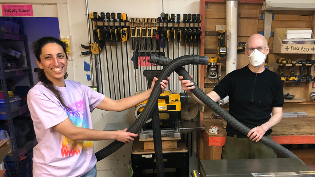 Two people holding vacuum hoses in woodworking workshop.